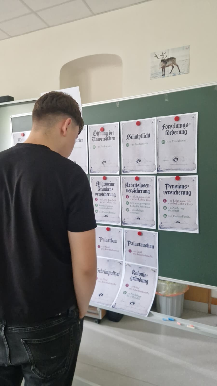 Schüler betrachtet an der Tafel Karten zu Krankenversicherung, Arbeitslosenversicherung und Pensionsversicherung